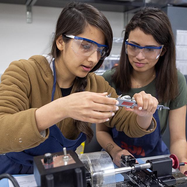 Engineering students carefully measure a machined part using a dial caliper